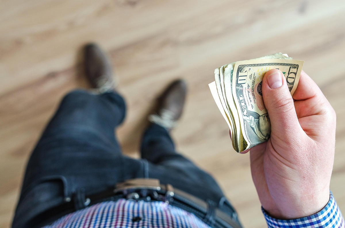 A pair of hands meticulously organizing stacks of various currency bills and coins on a modern wooden desk, with a partially visible digital budget spreadsheet on a tablet or laptop in the background, conveying careful financial planning and control.
